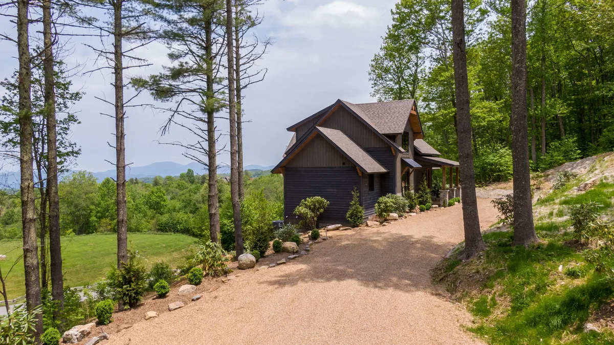 A wide shot of the retreat showcases the house set amid a serene landscape of trees and distant mountains. A gravel driveway leads up to the inviting porch, suggesting a peaceful, secluded getaway with natural beauty all around.