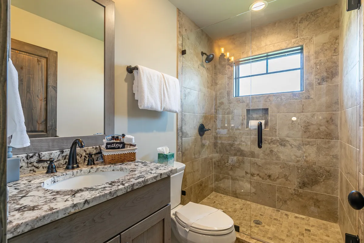 A modern bathroom with a granite countertop, double sinks, glass shower enclosure, wooden cabinetry, and warm lighting. The atmosphere is spa-like and serene, suggesting a place for rejuvenation after adventurous days.