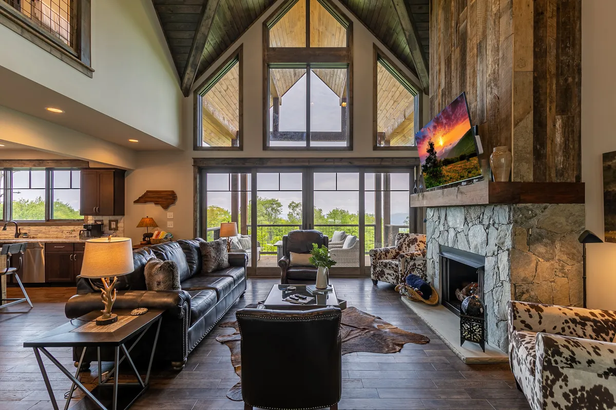 A cozy great room with vaulted ceilings, exposed wooden beams, a striking stone fireplace, and comfortable seating arranged to face the mountain view through tall windows. The space is filled with natural light and exudes warmth and luxury.