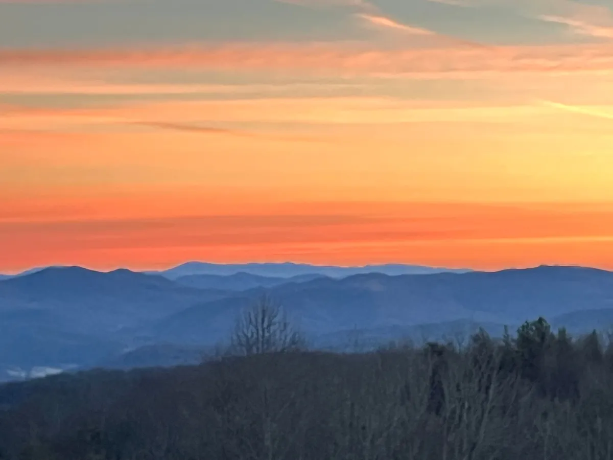 A breathtaking panoramic view of distant mountains at sunset, with silhouettes of trees in the foreground. The sky is vibrant with sunset hues, inviting feelings of serenity and awe, and suggesting the natural beauty that surrounds the property.
