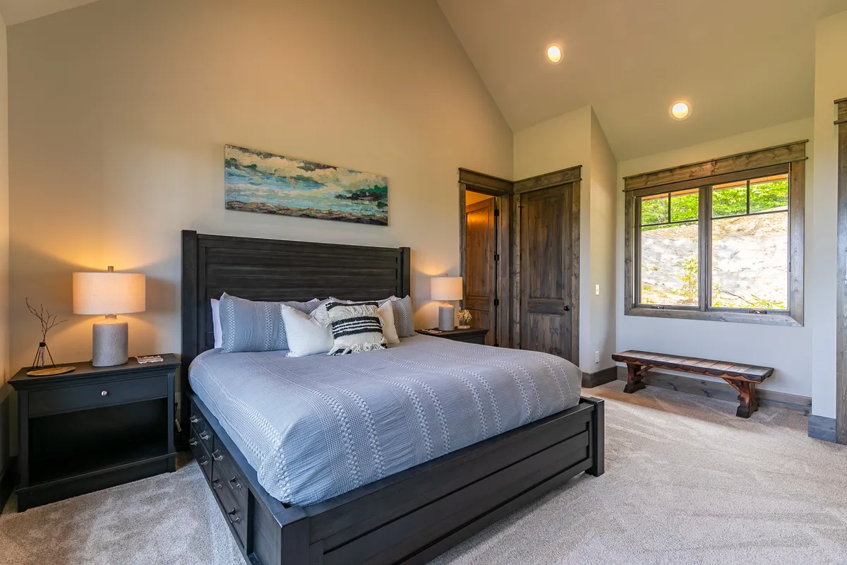 A serene and spacious bedroom featuring a king-sized bed with plush linens, a painting above the bed, a nightstand with a lamp, a window letting in abundant natural light, and a wooden bench at the foot of the bed. The overall atmosphere is tranquil, luxurious, and restful—perfect for couples or guests seeking a private retreat.