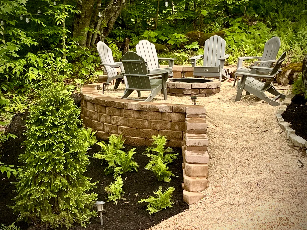An inviting exterior view of a fire pit surrounded by Adirondack chairs and lush greenery, suggesting a tranquil and social space for outdoor relaxation and evening gatherings.