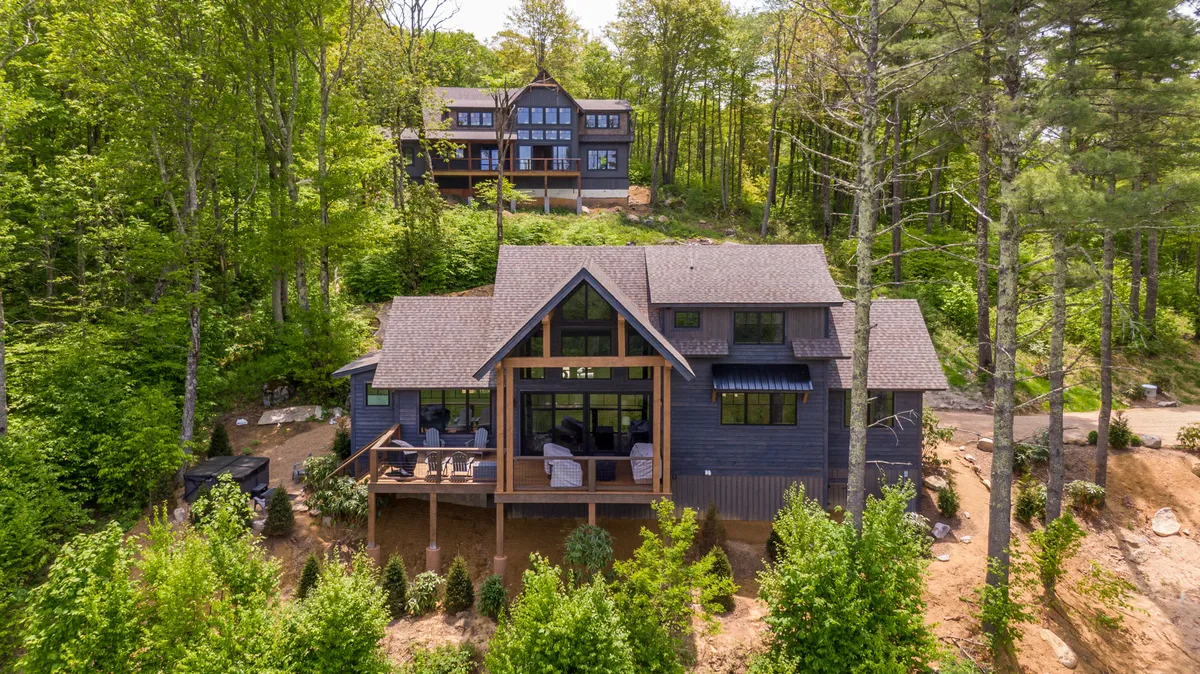A modern cabin with a spacious deck surrounded by tall trees. The deck features comfortable seating and expansive mountain views, ideal for relaxing or hosting outdoor gatherings. The ambiance is serene, with sunset lighting enhancing the inviting atmosphere.