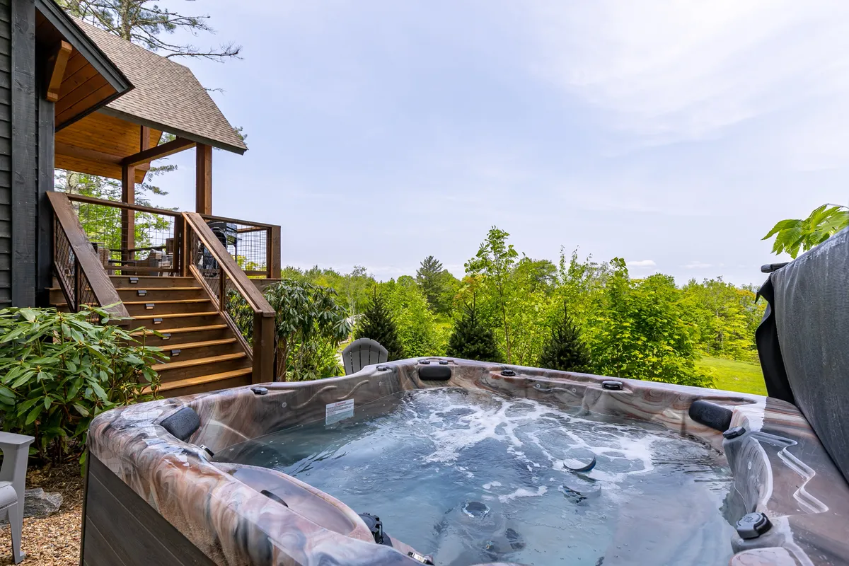 A wooden hot tub sits on a deck, accessed by nearby stairs and nestled amidst lush greenery. The setting is peaceful, perfect for socializing or relaxing in the bubbling water while enjoying the outdoors.
