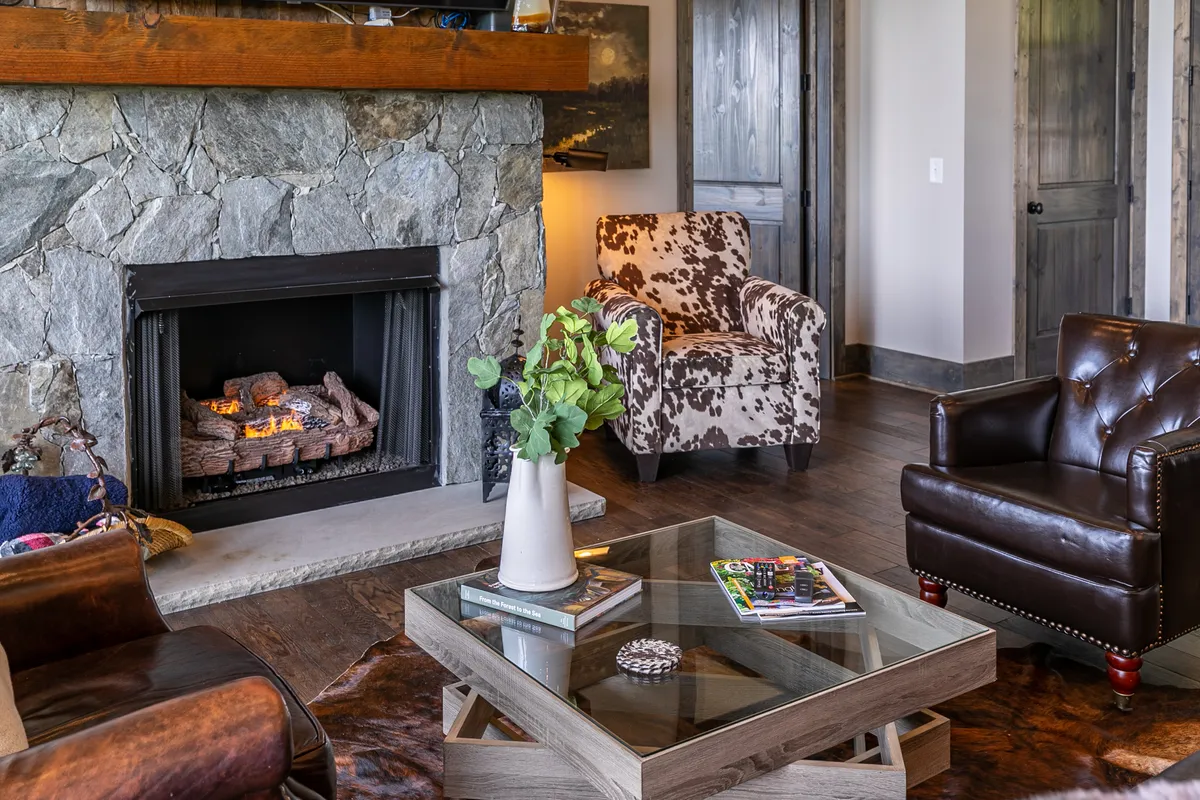 This image features a warm, inviting living room centered around a substantial stone fireplace, with a glass coffee table and plush cowhide-patterned and leather armchairs. The ambiance suggests a perfect setting for relaxing after outdoor adventures, particularly in winter or during chilly autumn evenings.
