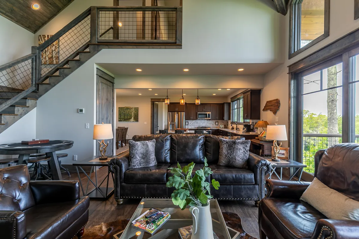 A wide-angle view of an open-concept great room featuring plush leather sofas arranged for conversation, a dining area set for eight, and a modern kitchen with granite countertops and stainless appliances. Large windows offer scenic views and flood the entire space with natural light.