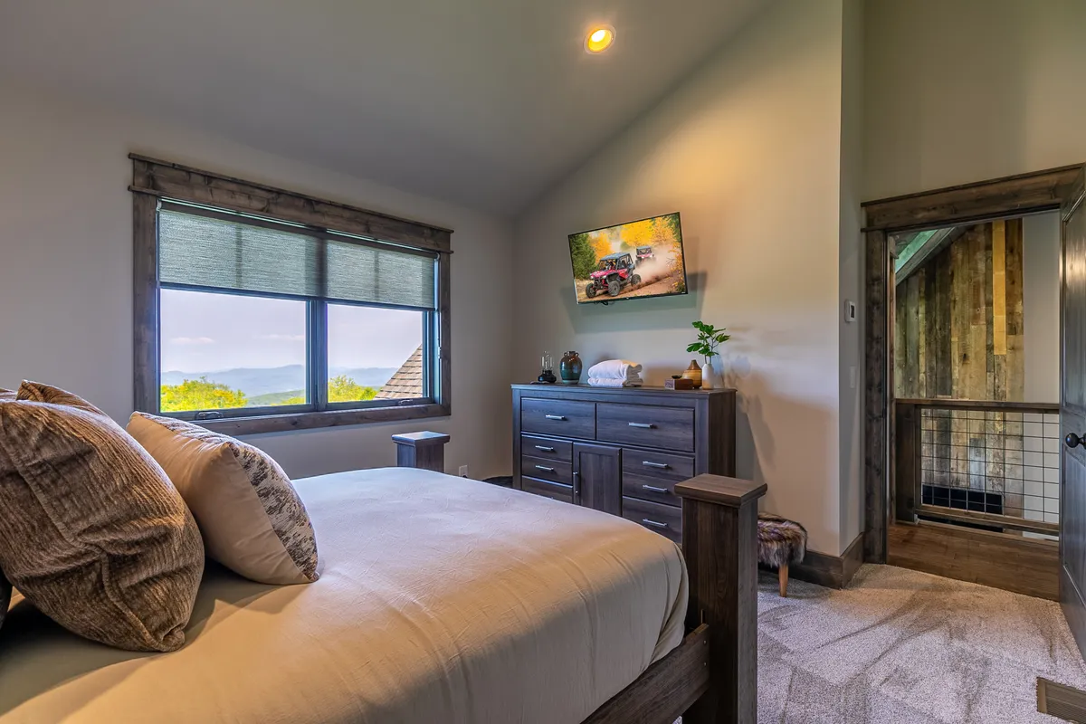 A queen bedroom with a cozy bed, window view, dresser, and a TV for entertainment. The room is styled in a comfortable and serene manner, ideal for guests who want both relaxation and convenience. Natural light filters in, enhancing the inviting mood.
