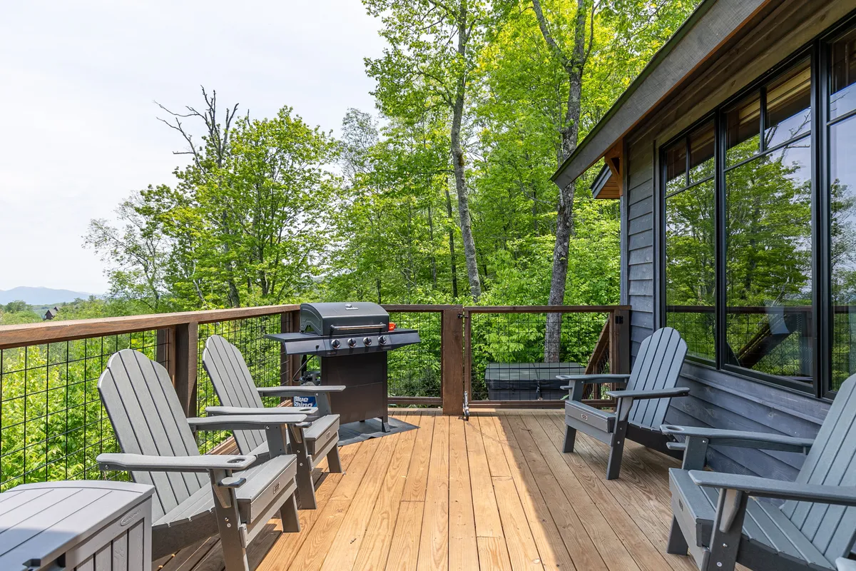 A set of Adirondack chairs and a gas grill are positioned on a deck, framed by natural greenery and trees. The space invites barbecuing and leisurely gatherings, making it ideal for alfresco meals with friends and family.