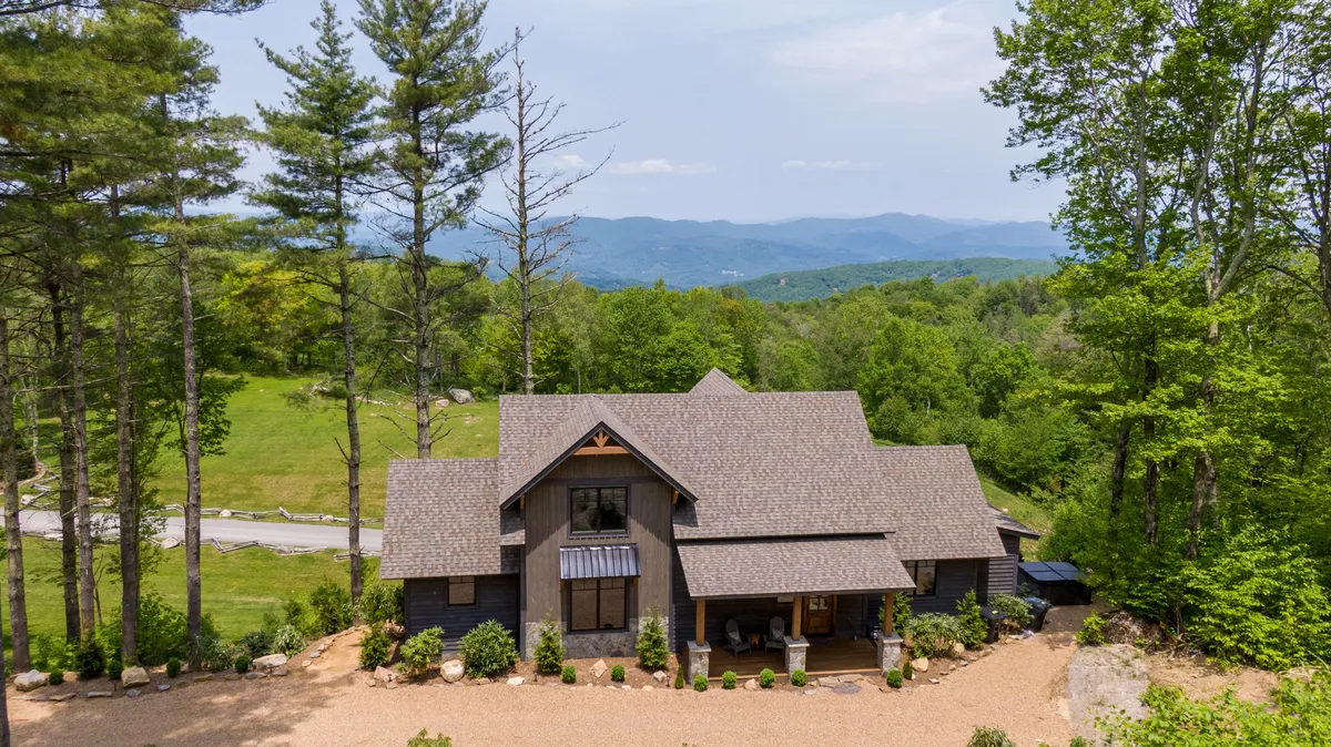 The image showcases a charming, newly built mountain cabin surrounded by lush trees and perfectly manicured gardens, perched atop a scenic bluff. In the foreground, the inviting porch offers sweeping sunrise views over the Blue Ridge Mountains, while the backdrop reveals endless skies and tranquil natural beauty. The setting radiates serenity, with ample outdoor space for gatherings, relaxation, and exploration.